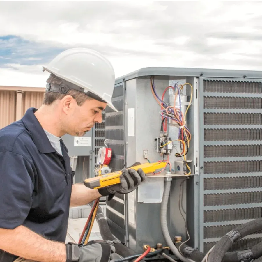 HVAC technician in a hard hat testing an outdoor air conditioner with a multimeter